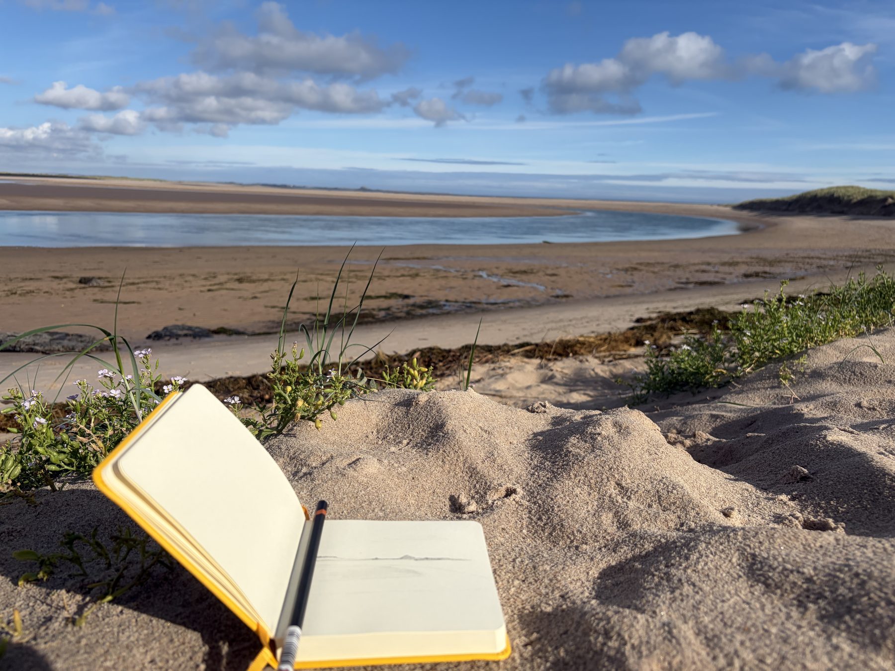 An open sketchbook and pencil resting in the sand, with the tide and dunes of Budle Bay beyond.